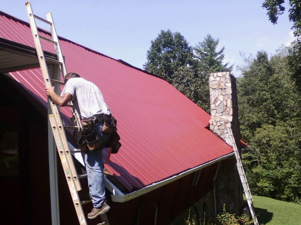 Roofer on a ladder beside a red metal roof and stone chimney — Soffit & Fascia Repair by Roofing Brothers in New Vienna